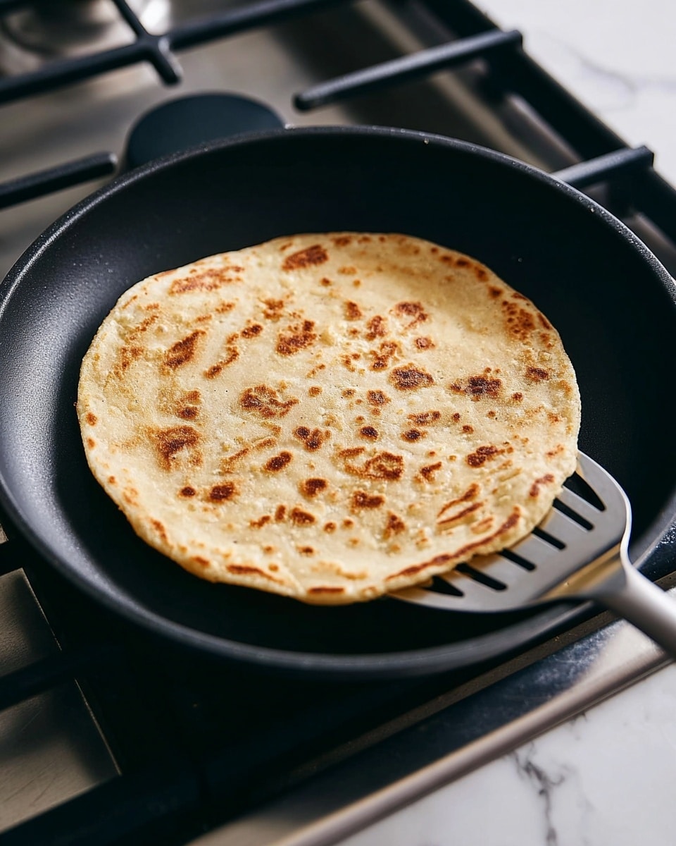 A single light brown flatbread with uneven golden toasted spots is cooking in a black frying pan on a silver stove. The flatbread is round and thin with a slightly rough texture, showing darker toasted patches scattered across its surface. The frying pan has a smooth, matte black interior and is being gently lifted at the edge by a metal spatula with slits, revealing the flatbread’s underside. The stove under the pan has a metallic finish with black burner grates. The background is a white marbled texture. photo taken with an iphone --ar 4:5 --v 7