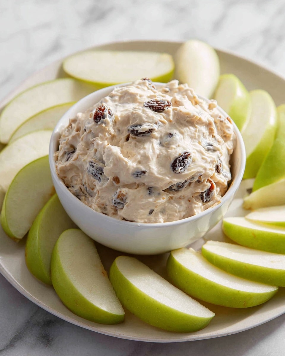 A small white bowl is filled with a creamy, light beige dip mixed with visible dark raisins and small crunchy pieces, giving it a textured and thick appearance. This bowl sits on a white plate surrounded by fresh, bright green apple slices with a smooth, shiny skin and white inner flesh. The background features a white marbled surface that adds a clean and fresh look to the overall image. photo taken with an iphone --ar 4:5 --v 7
