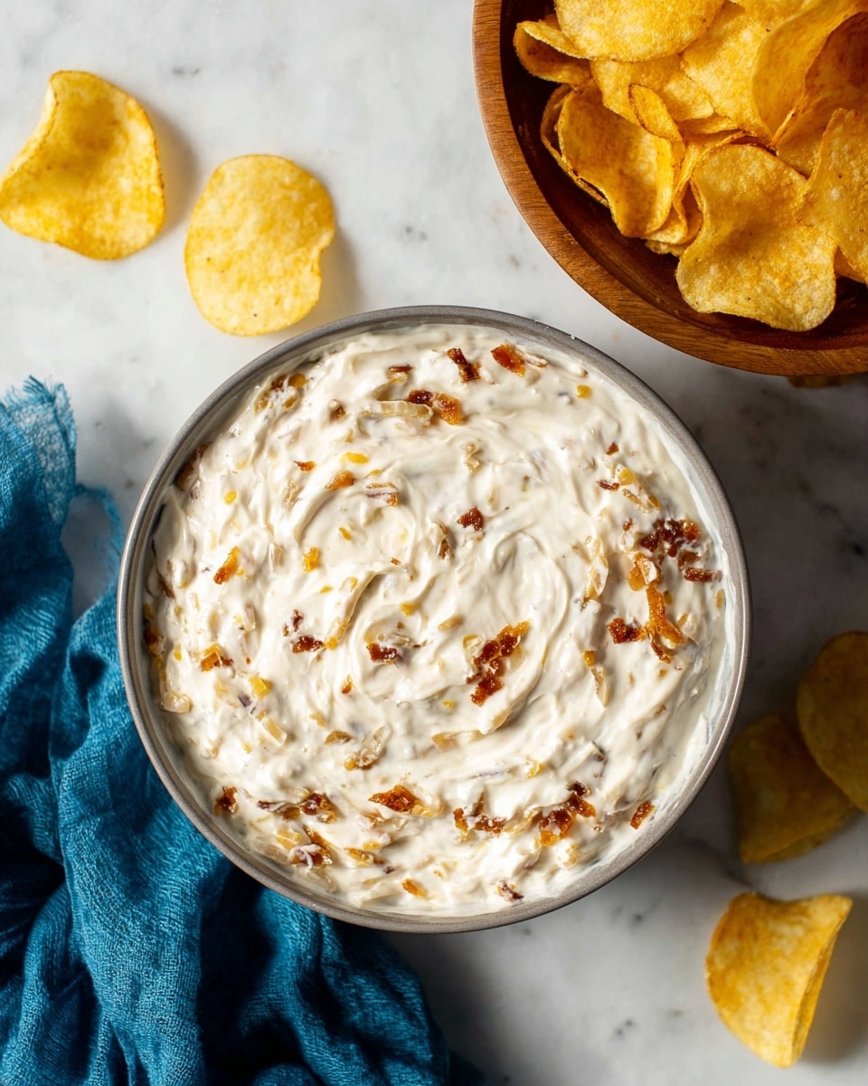 A close-up top view of a grey bowl filled with creamy white dip mixed with small pieces of golden brown toasted onions or bacon bits, giving the dip a textured look. The dip swirls smoothly inside the bowl with some peaks and valleys. To the right, there is a wooden bowl nearly full of golden yellow potato chips. Three potato chips are scattered on a white marbled surface near the bowls, and a crumpled blue cloth is placed towards the bottom left corner. The image has soft natural lighting highlighting the creamy texture of the dip and the crispy chips. photo taken with an iphone --ar 4:5 --v 7