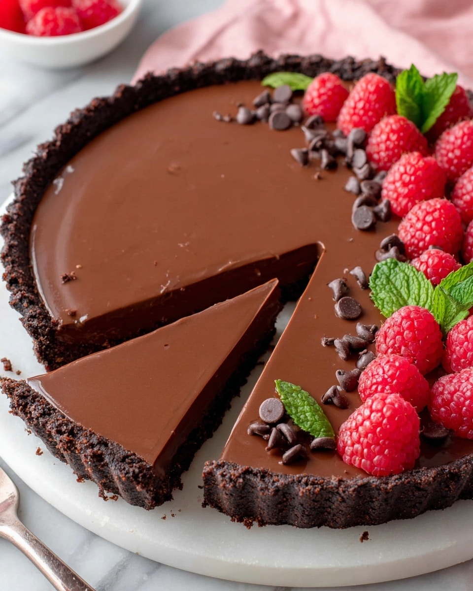 The image shows a chocolate tart with three distinct layers. The outer crust is dark brown and crumbly, forming a thick edge around the tart. The middle layer is smooth, shiny, and rich dark brown chocolate filling covering the entire tart surface evenly. On top of the chocolate layer, there is a decoration near the edge made of bright red raspberries, fresh green mint leaves, and scattered small, dark brown chocolate chips. The tart is on a white plate placed on a white marbled surface, with a pink cloth blurred in the background and a white bowl with chocolate chips to the side, photo taken with an iphone --ar 4:5 --v 7
