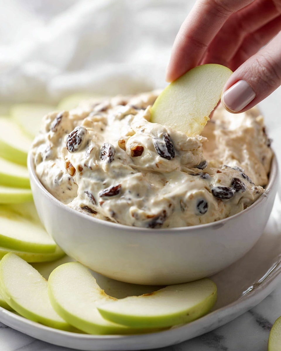 A close-up of a creamy dip with a thick, smooth texture mixed with dark small raisin-like bits and chopped light brown nuts, filling a white bowl that sits on a white plate. A woman's hand is dipping a crisp light green apple slice with a pale interior into the dip. Around the bowl, there are additional light green apple slices resting on a white marbled surface. The overall look is fresh and inviting. photo taken with an iphone --ar 4:5 --v 7