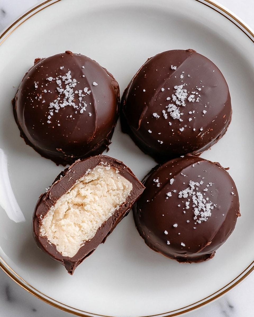 The image shows four round chocolate-covered candies on a white plate with a thin gold rim. Three of the candies are whole with a smooth, dark brown chocolate coating sprinkled with small white salt flakes on the top. The fourth candy is cut in half, revealing a thick, creamy, off-white filling inside with a rough texture. The chocolate layer is thin but clearly visible surrounding the filling. The plate is set on a white marbled surface. Photo taken with an iphone --ar 4:5 --v 7