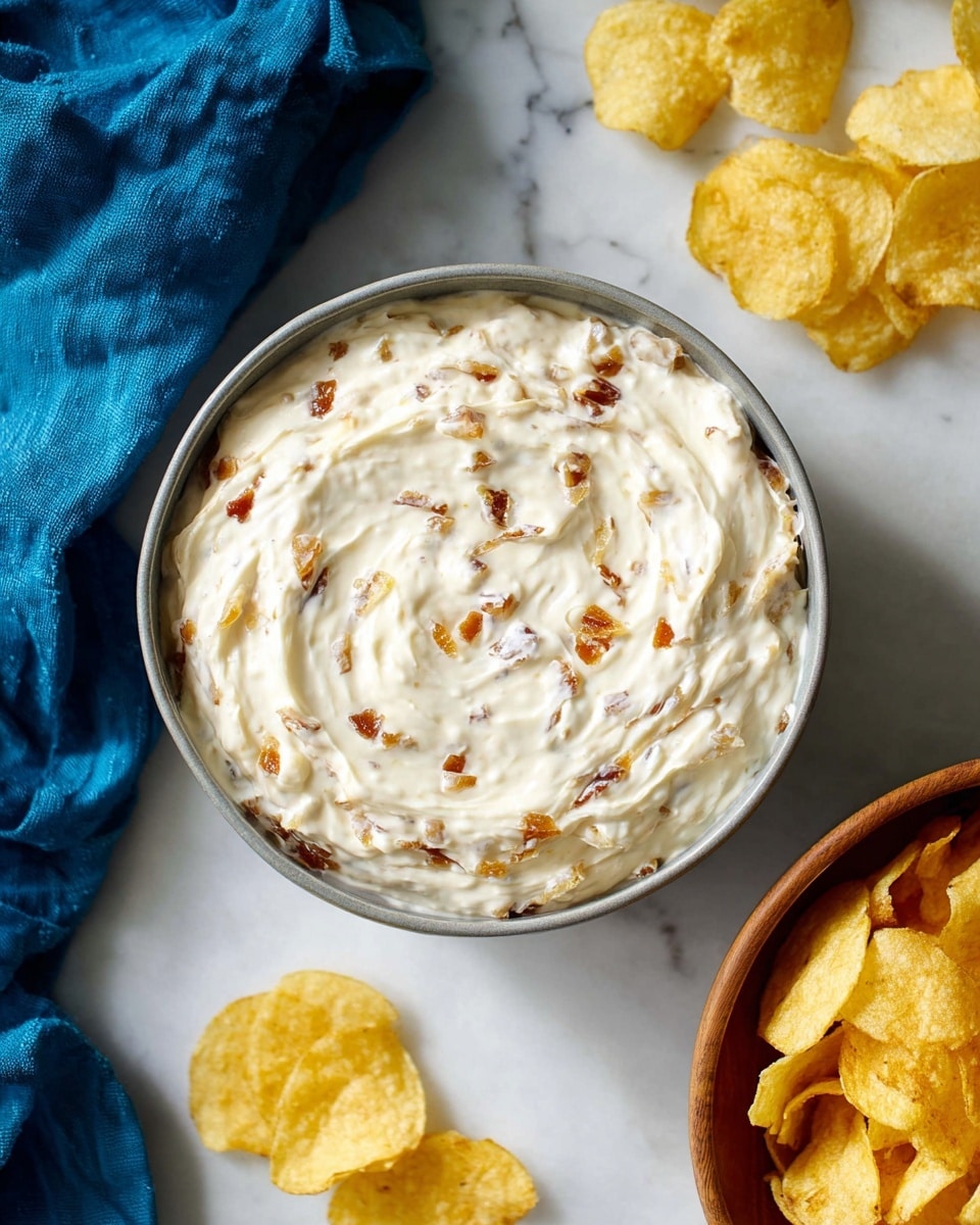 A top view of a grey bowl filled with creamy white dip mixed with light brown caramelized onion pieces throughout. The dip has a smooth and slightly chunky texture with swirls on the surface. To the right, there is a white bowl full of golden, slightly curled potato chips. A few potato chips are scattered on the white marbled surface around the bowls, and a blue cloth is placed near the bottom left corner. Photo taken with an iphone --ar 4:5 --v 7