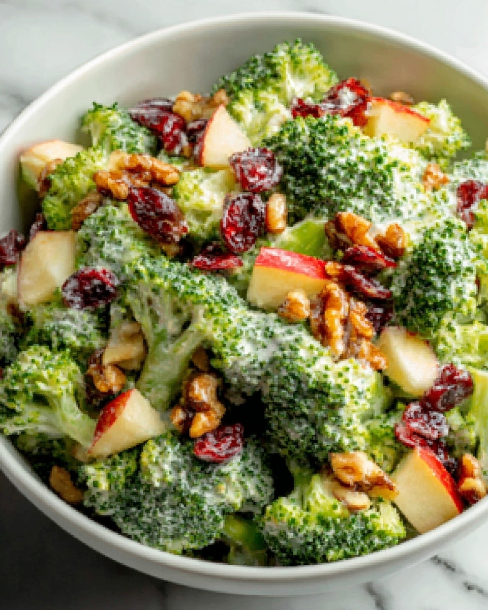 A white bowl filled with a broccoli salad sitting on a white marbled surface. The salad has several layers including bright green broccoli florets, light green creamy dressing covering the broccoli, small red dried cranberries scattered evenly, diced pale orange apples with some red skin pieces, and small brown walnut pieces spread over the top. The textures vary from soft creamy to crunchy and fresh. Photo taken with an iphone --ar 4:5 --v 7