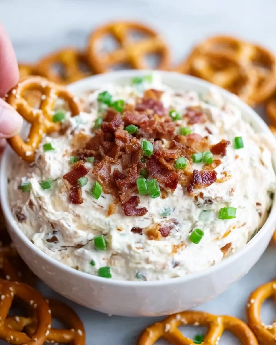 A white bowl filled with creamy white dip showing small bits of green and brown mixed inside. On top of the dip, there are crispy light brown bacon pieces and chopped green onion slices scattered. The bowl is on a white marbled surface, with parts of pretzels around it. A woman's hand is holding one pretzel close to the bowl. Photo taken with an iphone --ar 4:5 --v 7