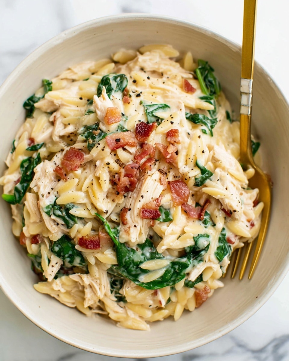In a close-up view, a white bowl holds a creamy pasta dish with three visible layers: the base layer of small, pale yellow orzo pasta, the middle layer of white shredded chicken, and the top layer of bright green fresh spinach leaves mixed throughout. Small bits of crispy, light brown bacon pieces are scattered evenly on and between the layers. The sauce coating the dish is smooth and creamy white, with flecks of black pepper sprinkled on top. A gold and white fork rests on the edge of the bowl, and the bowl sits against a white marbled surface. photo taken with an iphone --ar 4:5 --v 7