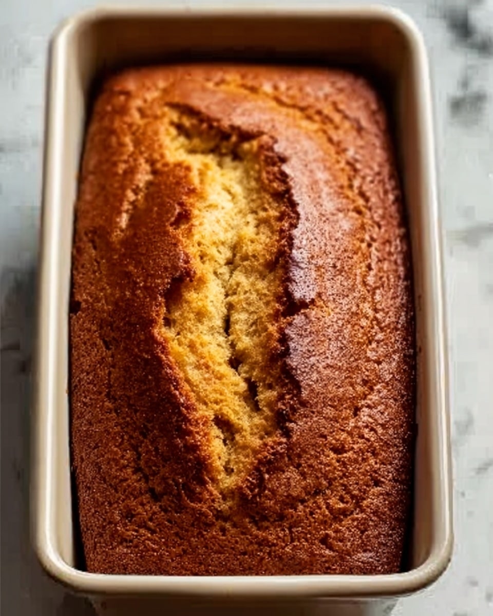 The image shows a freshly baked golden brown loaf cake in a white baking pan. The top of the cake has a cracked texture with a deep split running along the center, revealing a moist interior. The edges of the cake are slightly darker and crispy, while the middle is lighter and soft looking. The baking pan rests on a white marbled surface. The photo taken with an iphone --ar 4:5 --v 7