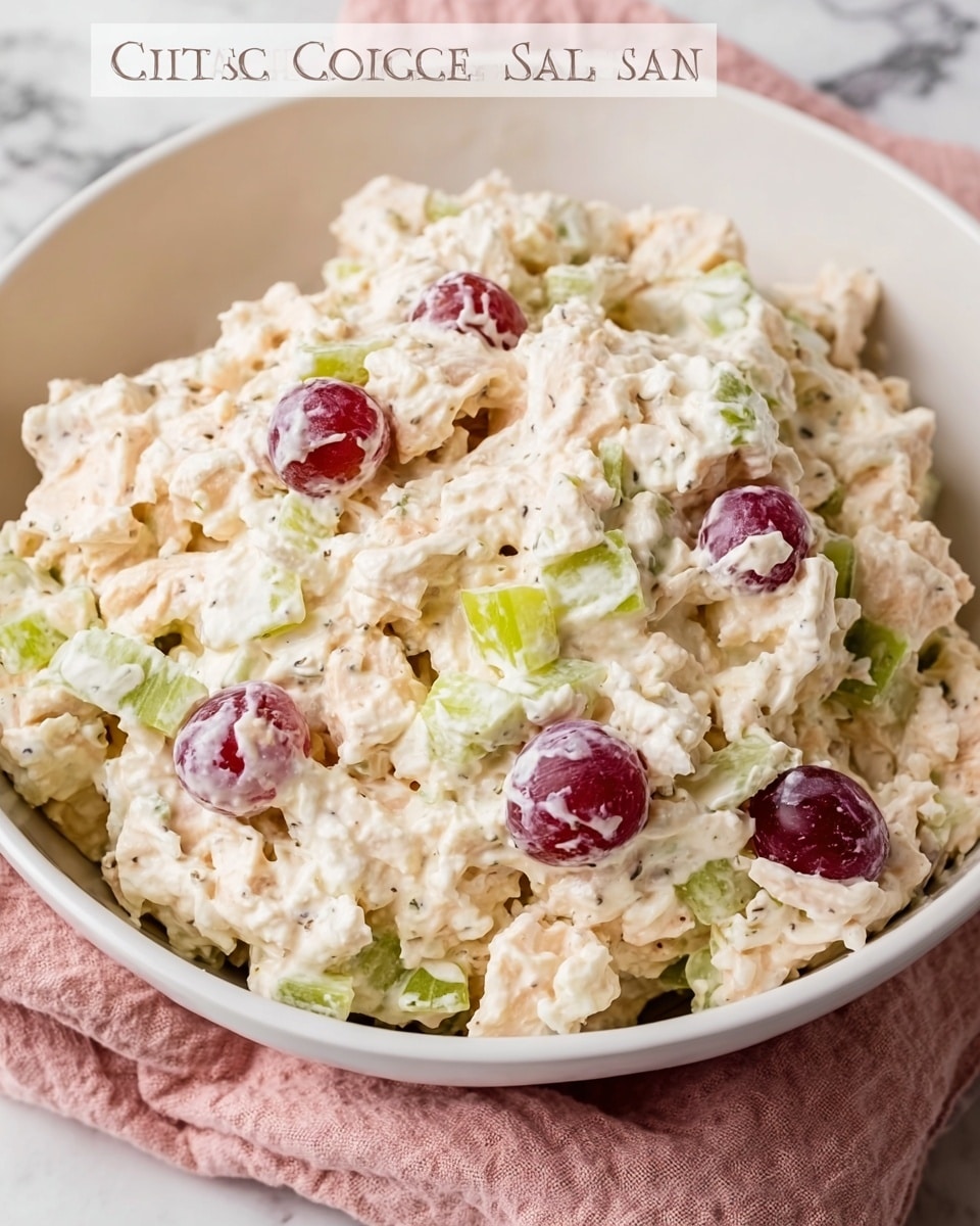 A white bowl filled with cottage cheese chicken salad showing a creamy, slightly chunky texture in off-white and light beige colors mixed with small pieces of green celery and red grape halves scattered throughout. The salad looks thick and rich, with visible specks of seasoning evenly spread. The bowl sits on a soft pink cloth against a white marbled surface, giving a fresh and clean look. Photo taken with an iphone --ar 4:5 --v 7