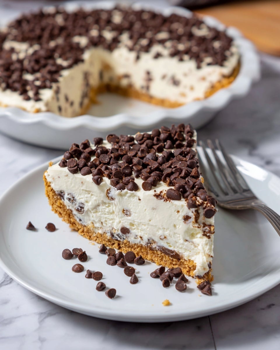 A slice of creamy pie sits on a white plate, showing three distinct layers: the bottom layer is a light brown crumbly crust, the thick middle layer is a white, fluffy filling mixed with small chocolate chips, and the top layer is a creamy white whipped topping covered densely with dark chocolate chips. Scattered chocolate chips lay around the slice on the plate, next to a shiny silver fork. In the background, more of the pie is seen in a white pie dish on a white marbled surface. photo taken with an iphone --ar 4:5 --v 7