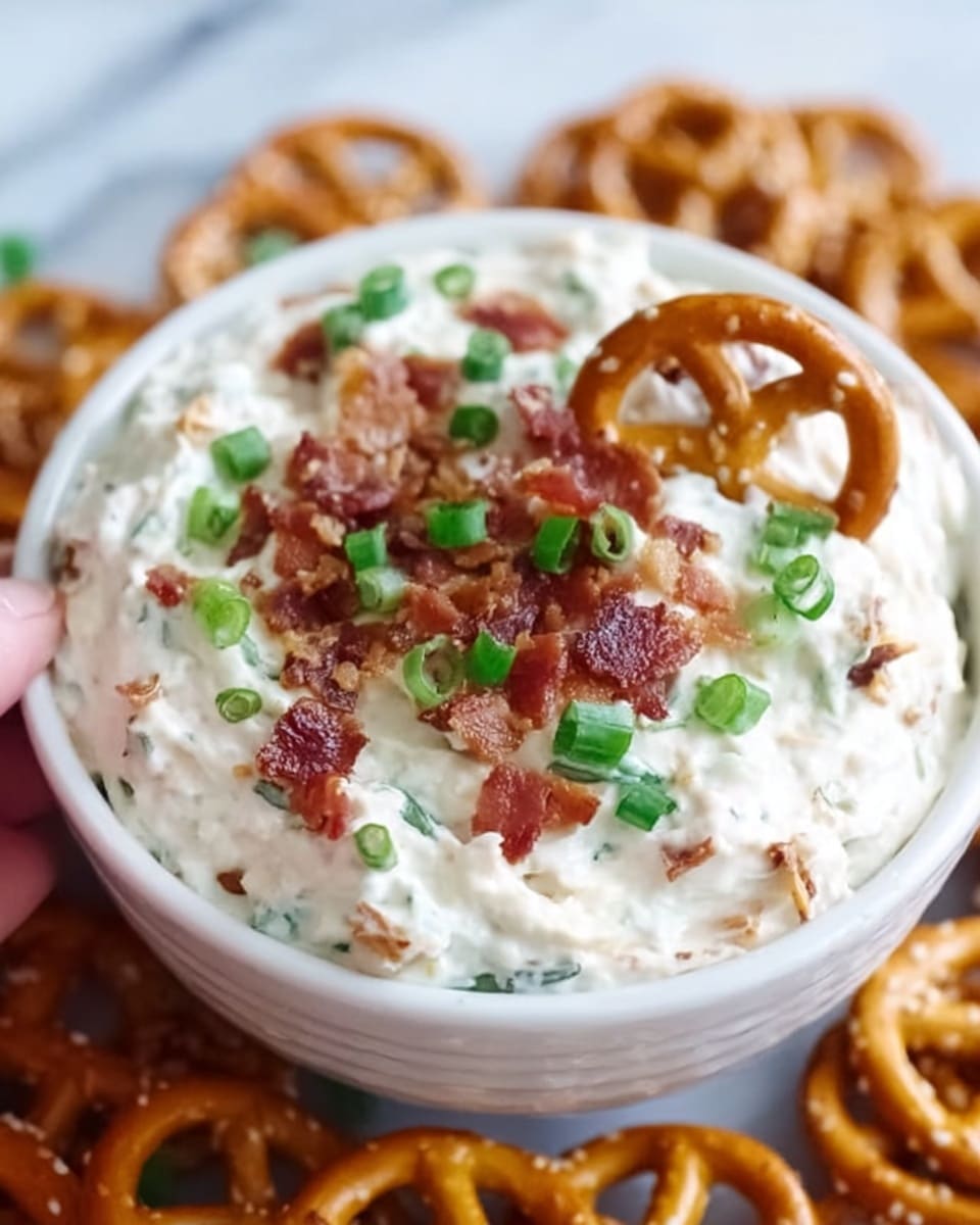 A white bowl filled with a creamy white dip that has a smooth, soft texture. On top, there are small green pieces of chopped onions and crispy brown bacon bits evenly spread, adding color contrast. The bowl sits on a white marbled surface, surrounded by light brown pretzels. A woman's hand is not visible but implied for serving. photo taken with an iphone --ar 4:5 --v 7