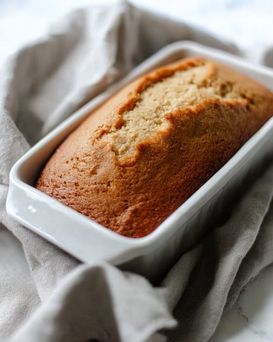 A close-up view of a golden brown loaf cake rising above the edges of a white rectangular baking dish. The cake has a slightly uneven, cracked top with a soft, fluffy texture. The dish sits on a light gray cloth with a slightly wrinkled texture, placed on a white marbled surface that fills the background with soft lighting creating a cozy atmosphere. photo taken with an iphone --ar 4:5 --v 7