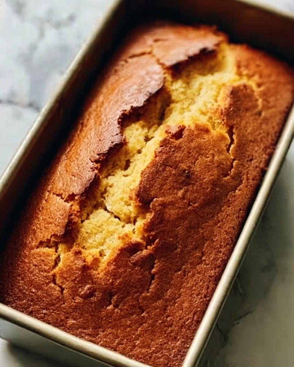 A close-up image of a golden brown loaf cake with a cracked top showing its soft inside, resting in a metal baking pan on a white marbled surface. The crust is slightly darker and textured with cracks running across it, exposing the moist, lighter-colored cake beneath. Light shines warmly on the cake, highlighting its baked, rustic texture. Photo taken with an iphone --ar 4:5 --v 7