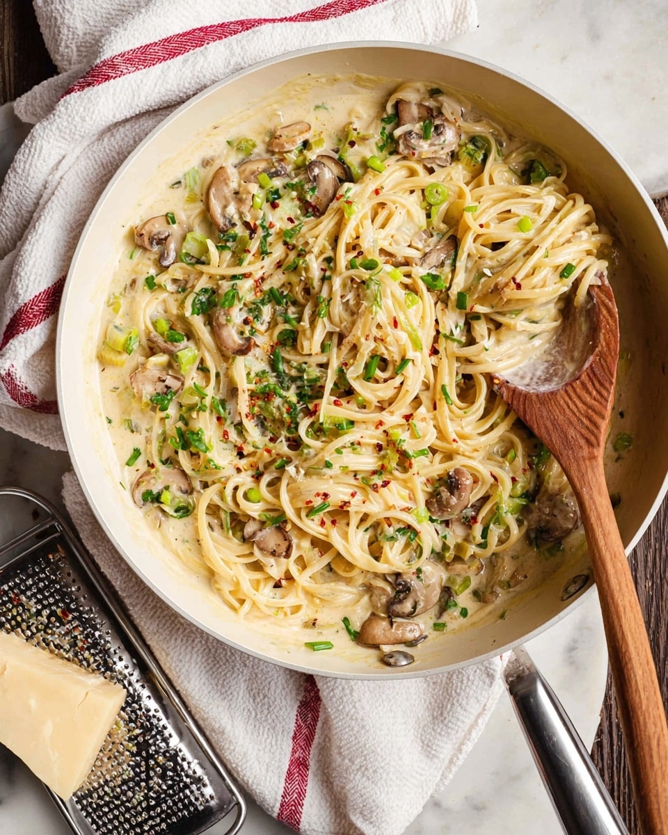 This image shows a close-up of a white pan filled with a creamy pasta dish. The dish has three main layers: the base layer is light cream sauce with a smooth texture, mixed evenly with thin, pale yellow linguine noodles that are soft and slightly curled. On top of the noodles, there are medium-sized mushroom pieces that are brownish-gray and look tender. These mushrooms are scattered evenly throughout the pasta. There are small bits of green herbs sprinkled on top, adding a fresh contrast. A wooden spoon is resting on the right side inside the pan, lifting some of the pasta. The pan sits on a wooden surface with a white towel with red stripes folded on the right side. At the bottom left corner, a block of light yellow parmesan cheese is placed on a white grater. The overall scene is warm and inviting, with natural lighting highlighting the pasta’s creamy texture. photo taken with an iphone --ar 4:5 --v 7