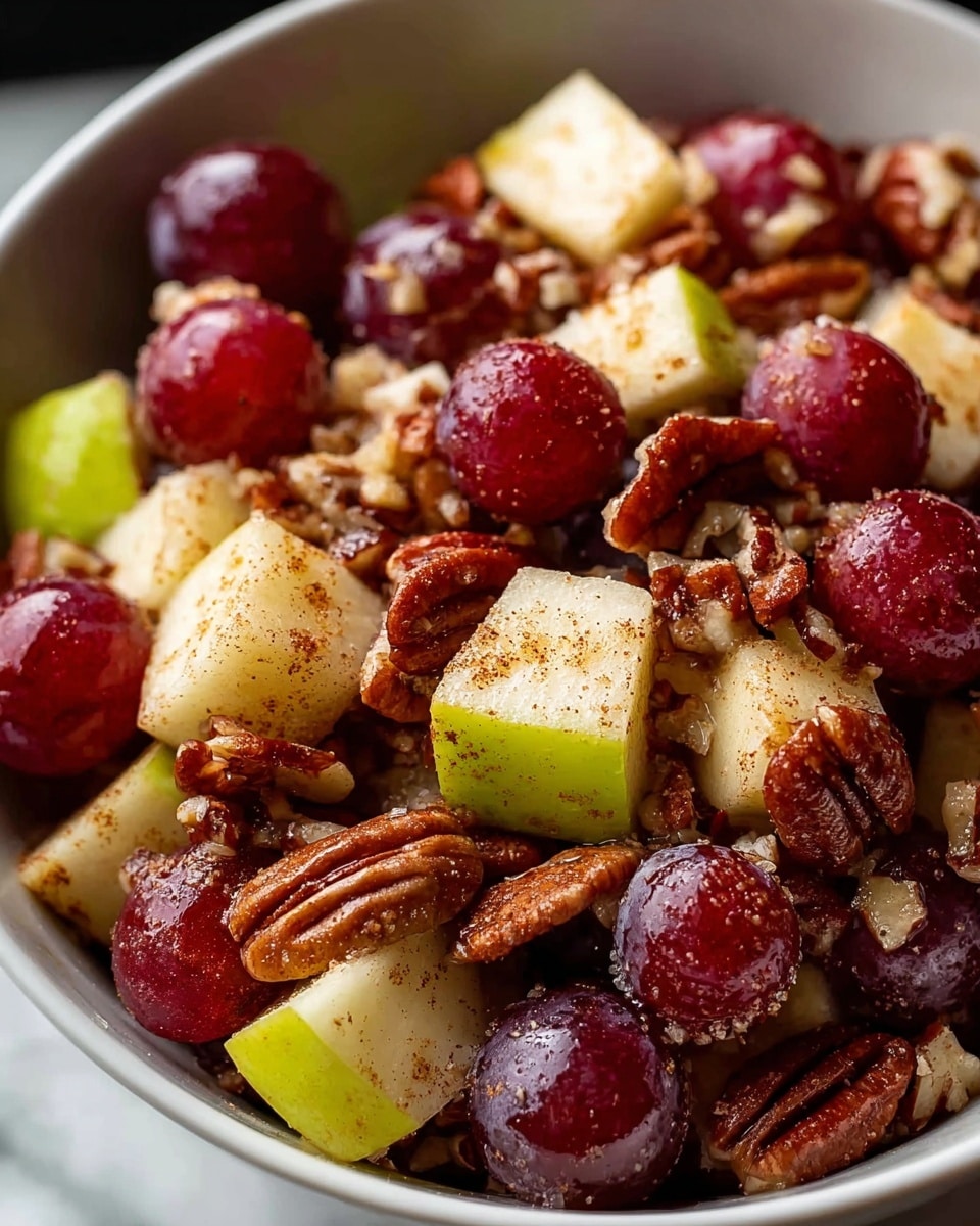 The image shows a close-up of a fruit and nut mix in a white bowl. The dish has three main layers visible: red grapes with smooth, shiny skins forming most of the top layer; chunks of pale yellow apple pieces with a slightly rough texture scattered evenly among the grapes; and whole pecan nuts with deep brown, ridged surfaces filling the spaces between the grapes and apple chunks. The nuts and fruit pieces are lightly coated with a brown spice dusting, giving the whole mix a textured and slightly glossy look. The background is a white marbled texture. photo taken with an iphone --ar 4:5 --v 7