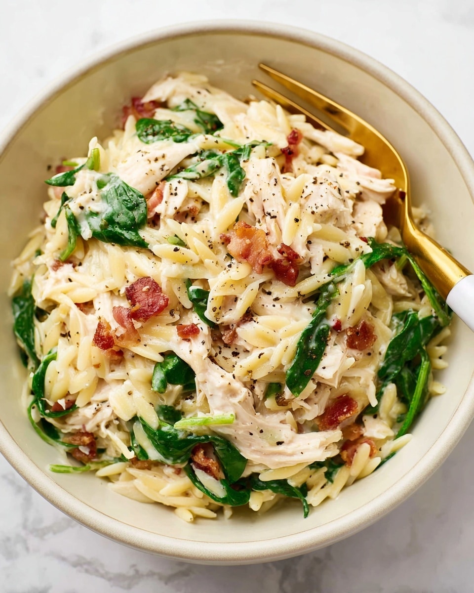 The image shows a close-up of a creamy pasta dish served in a white bowl. The dish has three main layers: al dente orzo pasta in a pale yellow color, tender shredded chicken in white, and fresh spinach leaves in bright green scattered through the dish. Light brown bits of crispy bacon or pancetta add small pops of color and texture throughout. The creamy sauce coating the pasta and chicken has a smooth, slightly glossy white appearance, with a few specks of black pepper sprinkled on top. A white and gold fork rests inside the bowl on the left side. The bowl sits on a white marbled surface. photo taken with an iphone --ar 4:5 --v 7