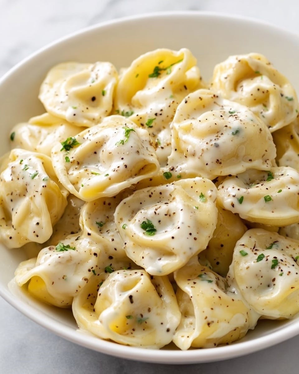A white plate filled with round, folded pasta pieces coated in a creamy white sauce with visible black pepper specks and small green herb bits sprinkled on top; the pasta looks soft and slightly glossy, stacked closely together with a smooth, rich texture. The background is a white marbled surface. Photo taken with an iphone --ar 4:5 --v 7