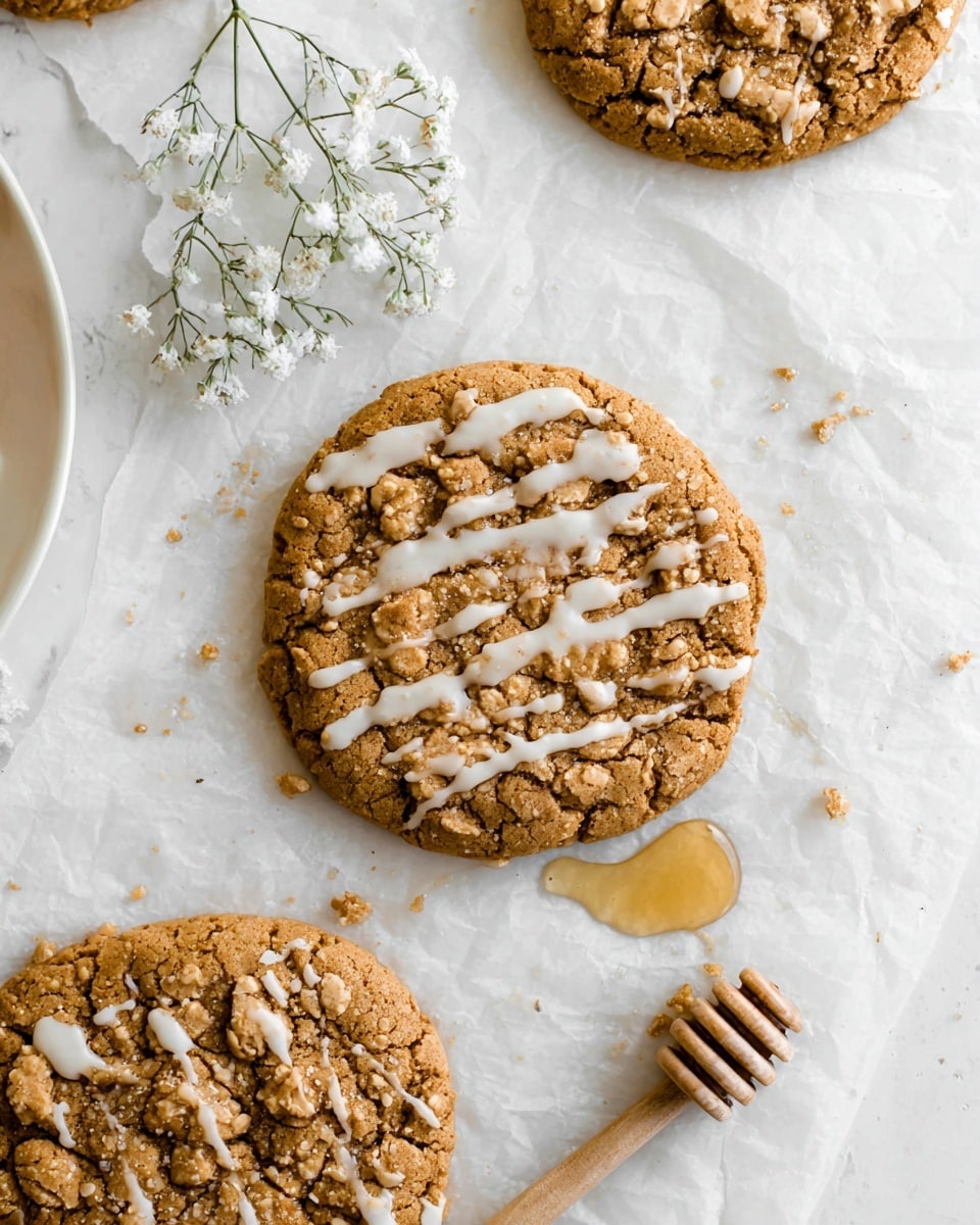 The image shows three large crumbly cookies on white parchment paper placed on a white marbled surface. The cookies are golden brown with a rough and crumbly texture on top; one in the center has a drizzle of white icing spread unevenly across its surface, creating thin lines and small drops around it. To the side, there is a small honey dipper with honey dripping near one cookie. A few delicate white baby's breath flowers are visible at the top, adding a light decorative touch. Part of a white plate with another cookie is seen on the left edge. Photo taken with an iphone --ar 4:5 --v 7