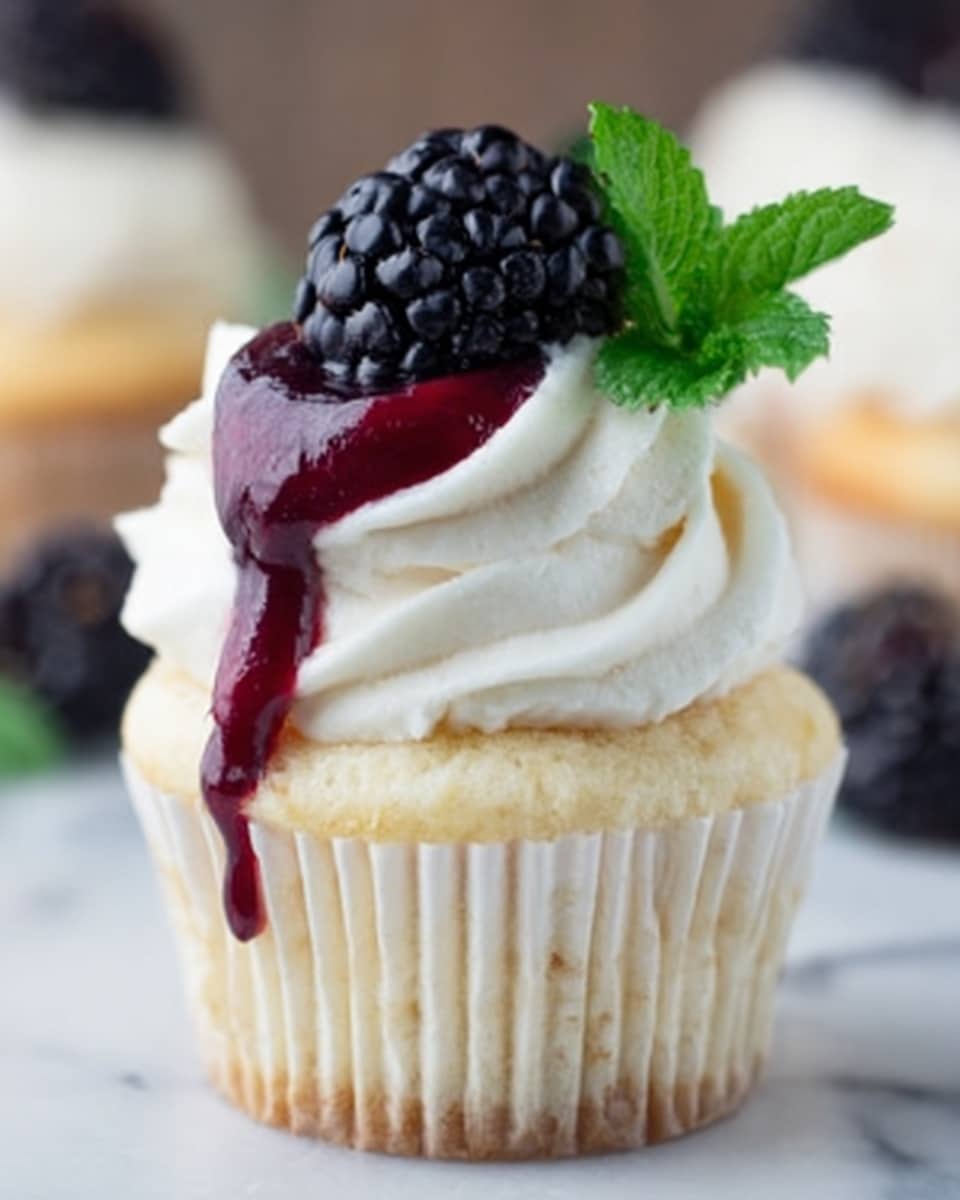 The image shows a single cupcake placed inside a white cupcake liner. The cupcake has three main layers: the base is a soft yellow cake, topped with a thick swirl of smooth white frosting. On top of the frosting sits a dark purple blackberry with a drizzle of glossy red berry sauce flowing down the frosting. A small green mint leaf is tucked beside the blackberry. The cupcake rests on a white marbled surface. photo taken with an iphone --ar 4:5 --v 7