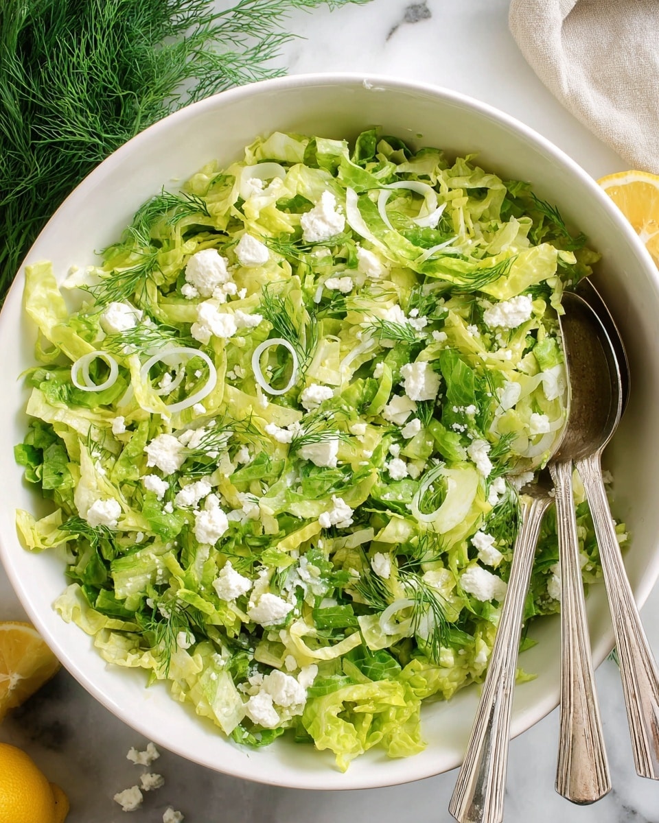 A white bowl filled with a fresh green salad made of shredded lettuce as the base layer, topped with thin slices of green onions, small fennel fronds, and crumbled white cheese scattered evenly over the top. The lettuce shows a mix of light and darker green shades with a crisp texture, while the white cheese adds a soft contrast. Two silver utensils, a fork and spoon, rest on the right side inside the bowl. The bowl is placed on a white marbled surface with some green herbs and lemon halves partially visible on the sides. Photo taken with an iphone --ar 4:5 --v 7