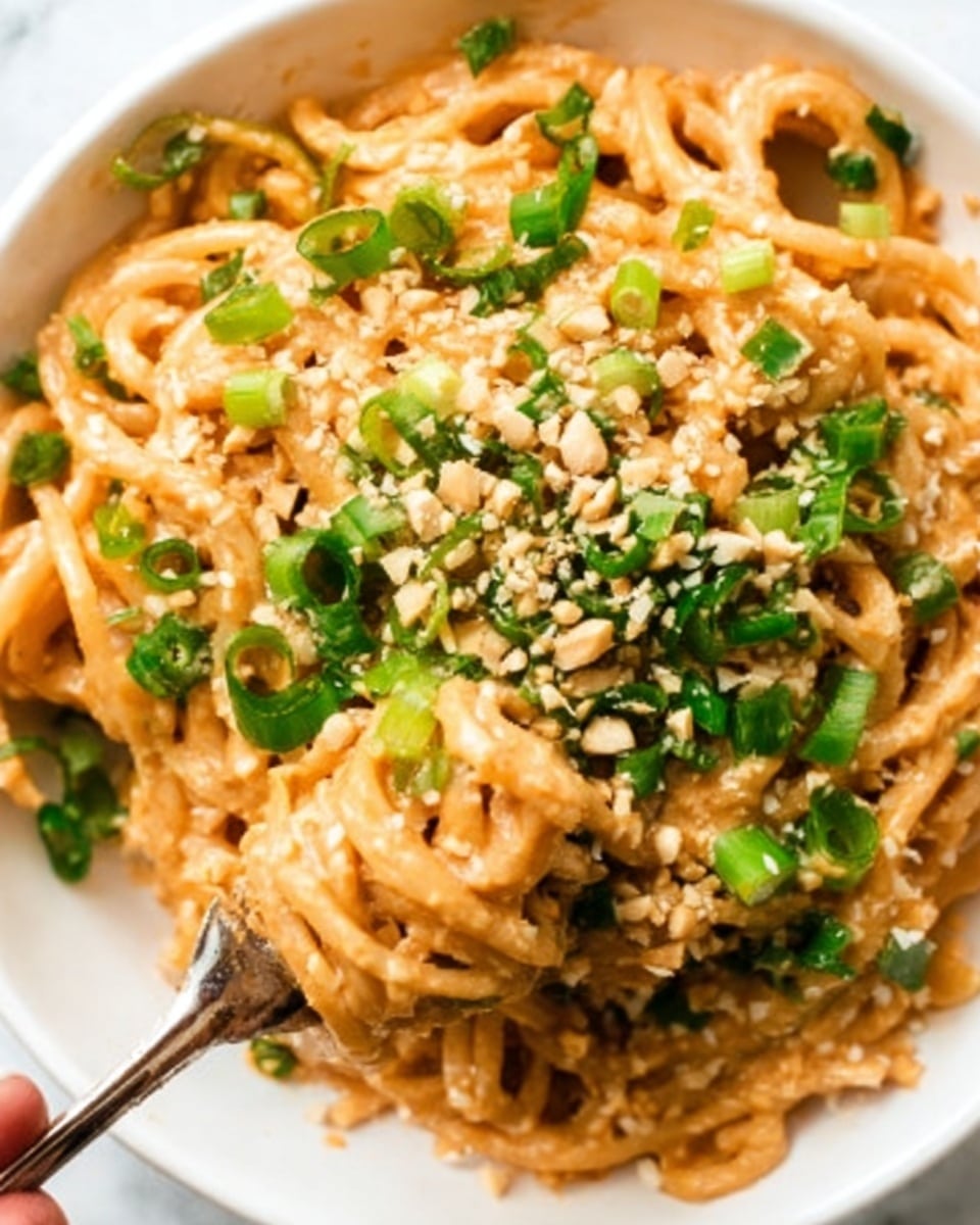 A close-up view of a bowl of noodles coated in a creamy peanut sauce, topped with chopped green onions, crushed peanuts, and sesame seeds, all arranged over the noodles. The thick noodles have a pale orange color from the sauce, with the green onions adding a vibrant green contrast on top. The crushed peanuts and sesame seeds give a textured, crunchy look over the soft noodles. The bowl is white, sitting on a white marbled surface, with a woman's hand holding a fork picking up some noodles from the side. Photo taken with an iphone --ar 4:5 --v 7