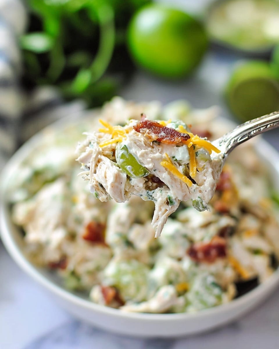 The image shows a close-up of a spoon holding a creamy chicken salad mix with visible chunks of white chicken meat, green celery pieces, small orange shredded cheese bits, and bits of crispy brown bacon. The background reveals a shallow white bowl filled with the same chicken salad, showing a mixture of white, green, orange, and brown colors with a soft texture. The scene is set on a white marbled surface with some blurred green leafy herbs and lime wedges surrounding the bowl, adding fresh color to the image. photo taken with an iphone --ar 4:5 --v 7