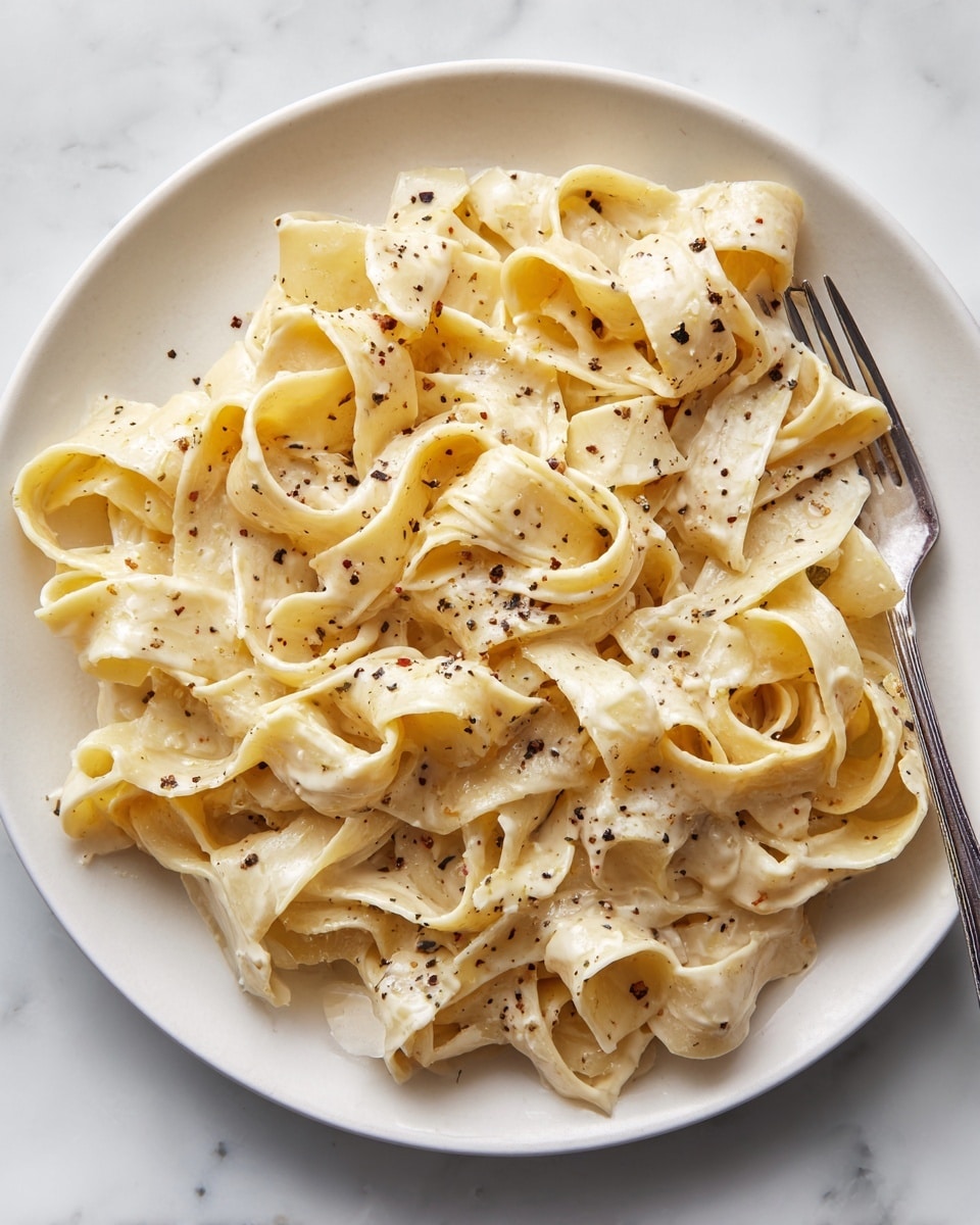 A close-up view of a white plate filled with wide, flat fettuccine pasta coated in a creamy light beige sauce. The noodles are layered in loose folds, glistening slightly with oil or butter, and sprinkled with small black pepper flakes that add contrast. A fork is resting on the right side of the plate, partially inserted under some pasta ribbons. The background features a white marbled texture. photo taken with an iphone --ar 4:5 --v 7