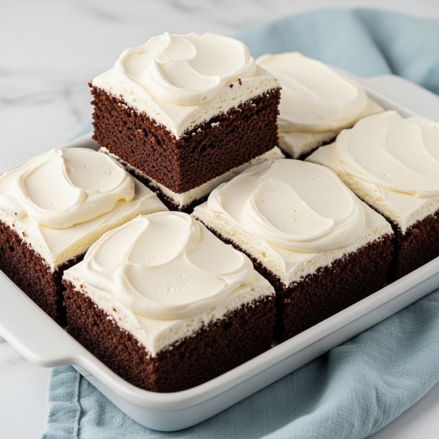 A close-up view of six square chocolate cakes in a white rectangular dish on a white marbled surface with a light blue cloth nearby. Each cake piece has a thick, soft layer of white creamy frosting spread unevenly on top with swirls and peaks. One piece is slightly raised above the others, showing its dark brown, moist and dense texture beneath the creamy frosting layer. The edges of the cakes are cleanly cut, highlighting their square shape. photo taken with an iphone --ar 4:5 --v 7