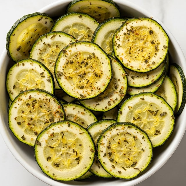 A close-up view of a white bowl filled with thin round slices of zucchini chips that are light green with darker green edges. Each chip shows a slightly crispy texture with specks of green herbs and light brown seasoning scattered across their surface. The chips are stacked loosely, some overlapping others inside the bowl. The background has a white marbled texture. photo taken with an iphone --ar 4:5 --v 7