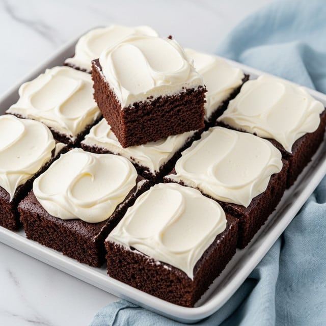 The image shows a white square baking dish filled with six thick, square pieces of chocolate cake topped with a generous layer of whipped cream. Each piece has a rich, dark brown, moist texture with a smooth, fluffy white layer of whipped cream evenly spread on top that contrasts with the deep color of the cake. One piece is lifted above the rest, showing its crumbly but moist chocolate texture clearly. The dish sits on a surface with a light green kitchen cloth beneath, all set on a white marbled texture. The whipped cream on each square has soft peaks and texture variations, making the dessert look fresh and inviting. Photo taken with an iphone --ar 4:5 --v 7