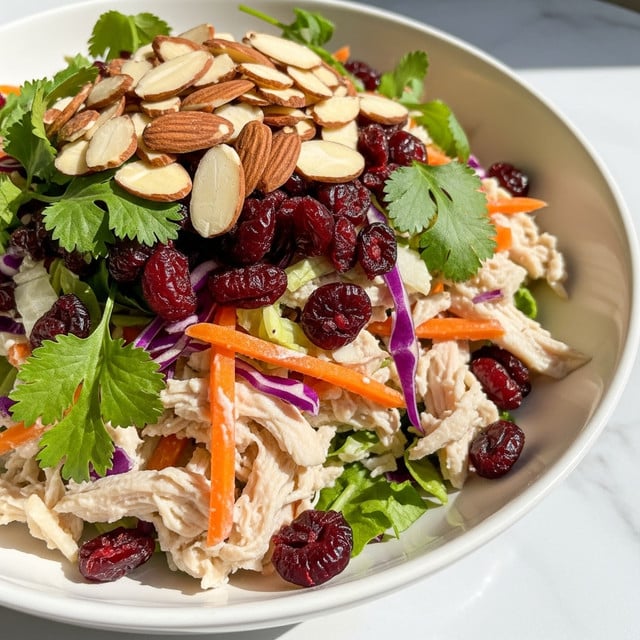 A white plate holds a colorful shredded salad made of light beige shredded chicken as the base layer mixed with thin orange carrot sticks and bits of light green cabbage. There are small bright purple cabbage pieces scattered throughout, adding deep color contrast. Dark red dried cranberries are spread evenly on top, bringing a shiny texture. The salad is garnished with light brown sliced almonds and bright green cilantro leaves scattered around, giving fresh touches. The plate sits on a white marbled surface with soft natural light shining on the salad. photo taken with an iphone --ar 4:5 --v 7