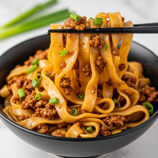 The dish shows wide, flat noodles coated in a shiny, brown sauce with small pieces of cooked ground meat mixed throughout. Bright green chopped scallions are sprinkled on top, adding a fresh touch. The noodles look soft and slightly translucent, being picked up with dark chopsticks above a deep, white bowl. The background includes a blurred bowl of green scallions, all set on a white marbled surface. photo taken with an iphone --ar 4:5 --v 7