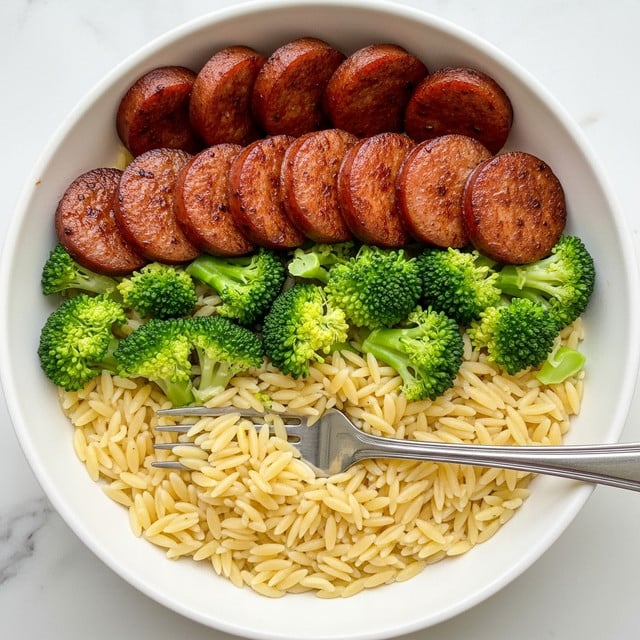 A white bowl filled with three main layers, with a silver fork resting inside. The bottom layer is small, light-yellow orzo pasta, soft in texture. The middle layer shows bright green broccoli florets scattered evenly around the bowl. On top, several thick slices of browned and crispy sausage sit, showing a mix of red and brown tones with a slightly charred surface. The ingredients are mixed but the layers remain distinct and colorful. The bowl is set on a white marbled surface. Photo taken with an iphone --ar 4:5 --v 7