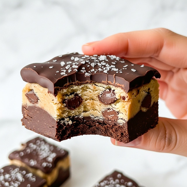 A close-up of a chocolate treat held by a woman's hand shows three main layers: a thick, smooth dark chocolate bottom layer with a matte finish, a thick middle layer of light tan cookie dough mixed with large, glossy dark chocolate chips of different sizes embedded inside, and a glossy dark chocolate top layer with an uneven surface sprinkled with coarse white sea salt crystals. The background has several similar chocolate pieces slightly blurred on a white marbled texture. photo taken with an iphone --ar 4:5 --v 7