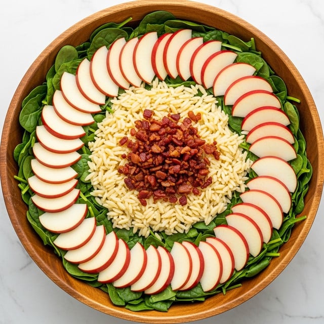 A large wooden bowl is filled with a colorful, fresh salad showing many layers and textures. The bottom layer has dark green leafy spinach, topped with small light yellow orzo pasta scattered throughout. On top of this are small chunks of bright red and pale yellow chopped apples, mixed with crispy reddish-brown bacon bits and sprinkled herbs or pepper. The salad looks fresh and vibrant with a mix of soft, crunchy, and chewy textures. The background shows a few whole apples on a white marbled surface. Photo taken with an iphone --ar 4:5 --v 7