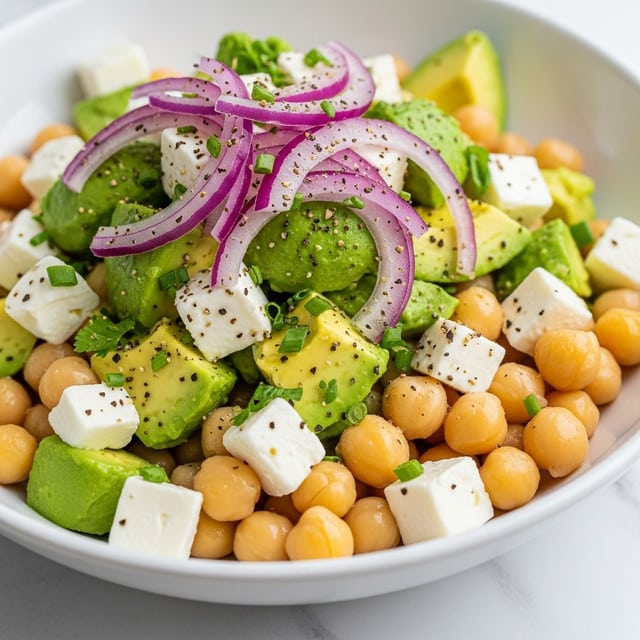 The dish is a bowl of salad with three main layers visible. The bottom layer has light beige chickpeas scattered evenly. The middle layer is made of bright green avocado pieces cut into small cubes with a smooth texture. On top, there are thin slices of purple-red onion placed randomly. Small white cubes of feta cheese are mixed throughout, adding a soft crumbly texture. The salad is sprinkled with finely chopped green herbs and a dusting of black pepper. The bowl is white, and it sits on a white marbled surface. Photo taken with an iphone --ar 4:5 --v 7