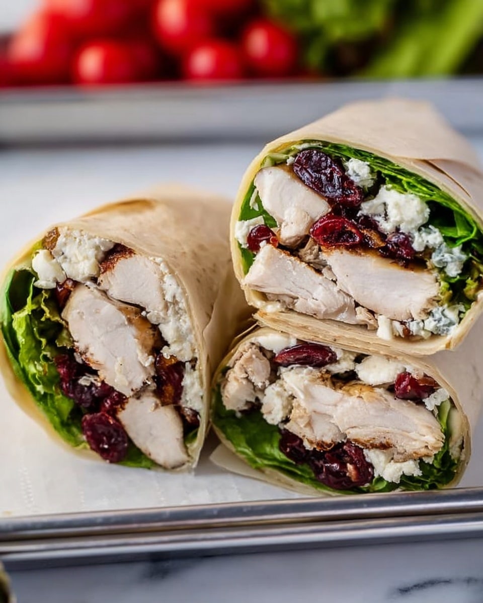 The image shows two halves of a wrap sandwich standing side by side. The wrap has multiple visible layers: the outer layer is a light beige tortilla, inside which there is a layer of fresh green leafy lettuce surrounding thick, juicy, white chicken pieces with a slight golden brown sear. Among the chicken, small dark red dried cranberries are spread throughout, along with crumbled white goat cheese that adds a soft texture. The wrap is placed on a metal tray, which rests on a white marbled surface. In the blurred background, red cherry tomatoes and some green vegetables can be seen. photo taken with an iphone --ar 4:5 --v 7