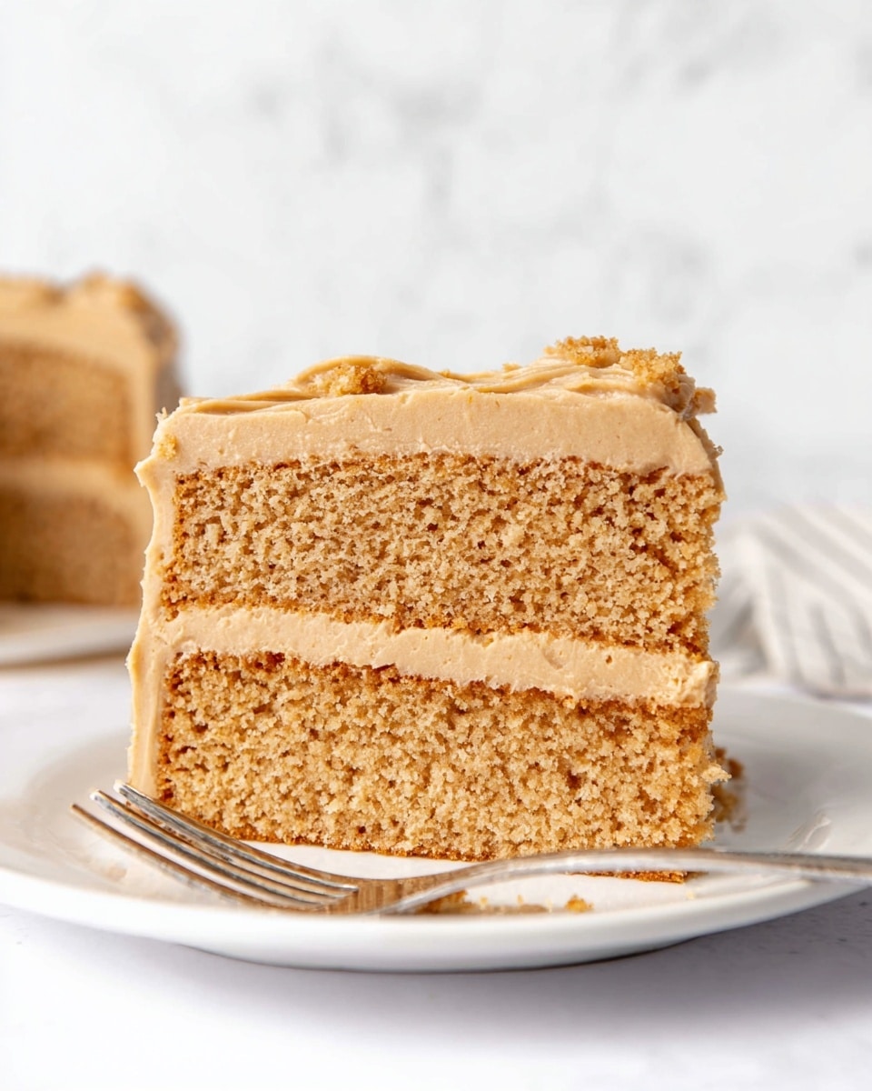 A slice of two-layer light brown cake with crumbly texture sits on a white plate, each layer separated by a thick layer of smooth, slightly darker brown frosting. The top layer is covered with the same frosting, which appears creamy and evenly spread, with small crumbs on the surface. The background is a white marbled texture, and a silver fork lies in front of the plate. photo taken with an iphone --ar 4:5 --v 7