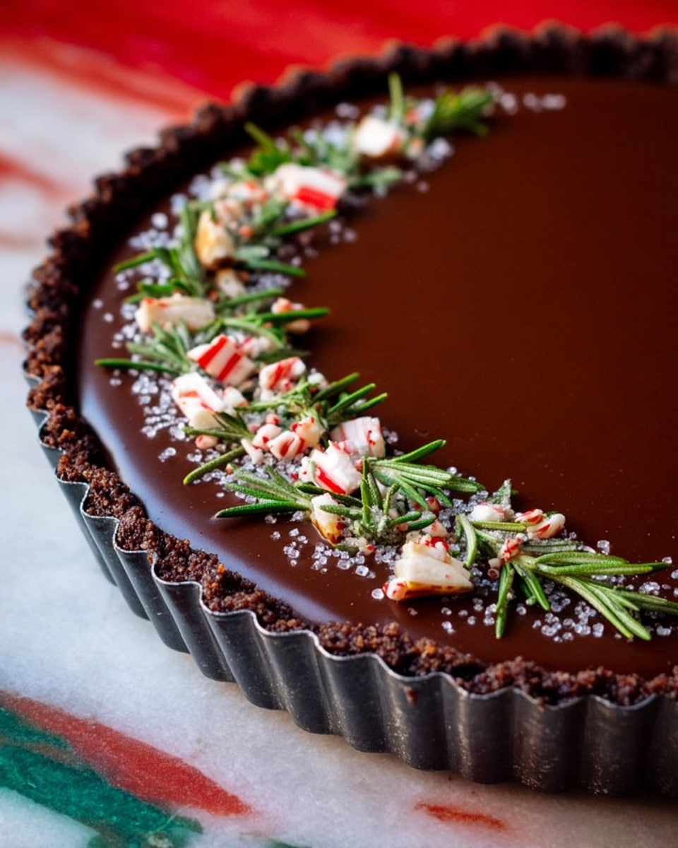 The image shows a chocolate tart with one smooth, shiny dark brown layer filling the tart shell. The tart shell is crumbly and dark brown, with a scalloped edge in a shiny metal tart pan. Across the top of the tart, from one side to the other, there is a line of decoration made of small green rosemary sprigs, broken pieces of red and white candy, and small white round sprinkles. The tart is placed on a white marbled surface with a red and green blurry pattern near the bottom left corner. photo taken with an iphone --ar 4:5 --v 7
