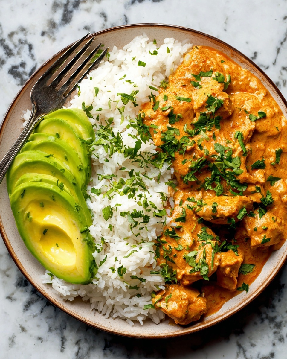 A white plate holds a serving of fluffy white rice as the base layer, sprinkled with chopped green herbs on top. On the right side, there is a layer of rich, creamy orange-colored chicken curry with visible chunks of chicken, also garnished with fresh green herbs. On the left side of the plate, there are neatly sliced pieces of bright green avocado arranged in a fan shape. A fork rests on the left side of the plate. The plate sits on a white marbled surface. Photo taken with an iphone --ar 4:5 --v 7