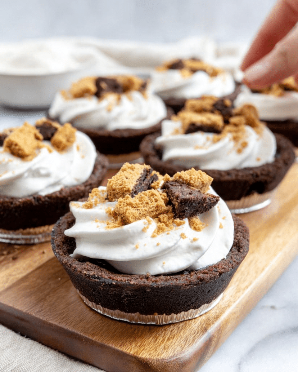 The image shows a close-up of small round chocolate pies in foil containers, each with one thick and slightly cracked dark brown cake layer as the base. On top of each cake layer is a swirl of smooth white cream arranged in a circular pattern near the edge, leaving the center open. The center is topped with golden brown cookie pieces that look crunchy and crumbly. The pies are set on a wooden board with a white marbled surface in the background, and a woman's hand is about to pick one pie from the board. Photo taken with an iphone --ar 4:5 --v 7