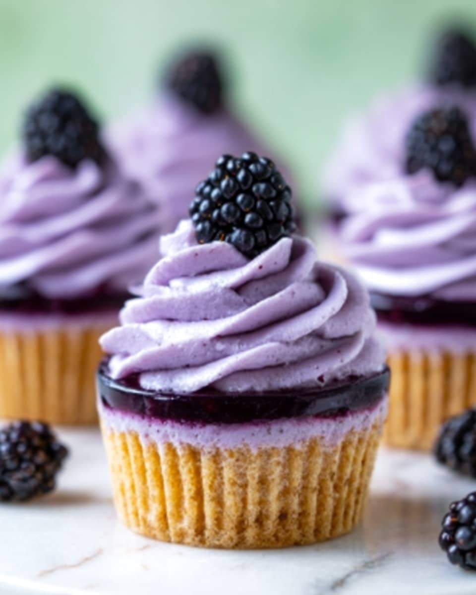 The image shows close-up of light purple cupcakes with swirled lavender frosting on top. Each cupcake has a dark purple layer of jelly just below the frosting, visible on the top surface. A single fresh blackberry sits on the peak of each frosting swirl. The cupcakes are placed on a white marbled surface with a soft green background that is out of focus. The texture of the frosting looks smooth and creamy, while the cupcake base appears light and fluffy. Photo taken with an iphone --ar 4:5 --v 7