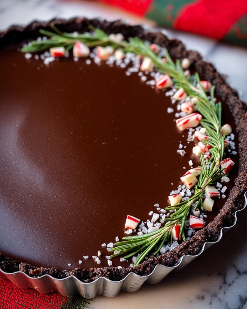 A chocolate tart with one thick dark brown shiny smooth layer of chocolate filling inside a crumbly dark chocolate crust, held in a silver tart pan with scalloped edges. On top, there is a neat line of small white sugar pearls, broken red and white candy pieces, and green rosemary sprigs arranged in a row from near to far edge. The tart is set on a white marbled textured surface with part of a red and green cloth visible at the side. Photo taken with an iphone --ar 4:5 --v 7