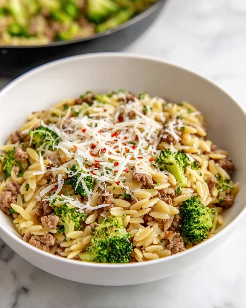 A white bowl filled with three main layers: the bottom layer consists of small green broccoli florets, the middle layer has cooked orzo pasta mixed with ground meat showing a light brown color and slightly crumbly texture, and the top layer is a sprinkle of white shredded cheese with a few red chili flakes scattered over it. The bowl sits on a white marbled surface and a frying pan with similar food is blurred in the background. photo taken with an iphone --ar 4:5 --v 7
