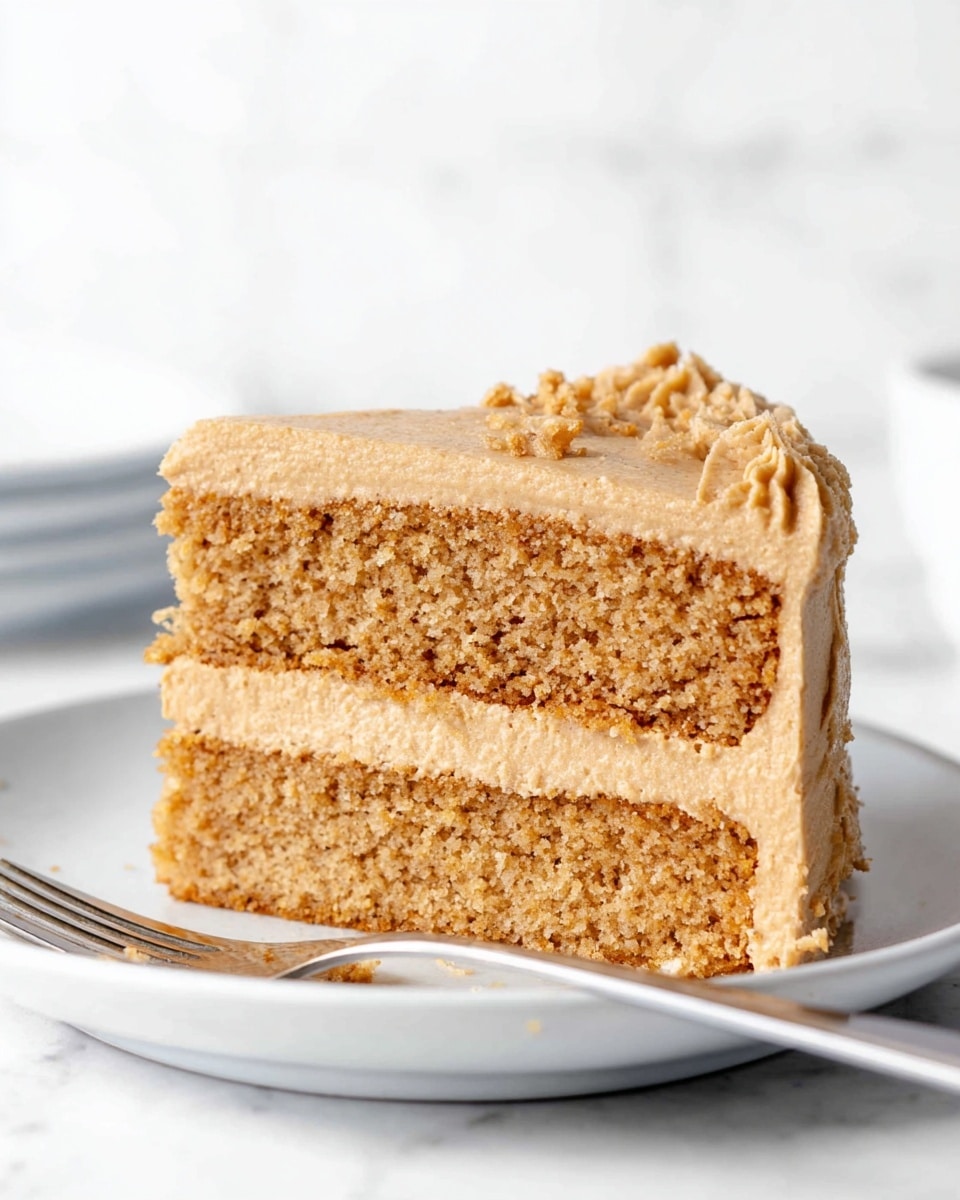 A close-up view of a two-layer cake slice on a white plate, placed on a white marbled surface. The bottom and top cake layers are light brown and moist with a crumbly texture. Between the two cake layers is a thick layer of creamy, tan frosting mixed with a crunchy, darker brown crumbly layer. The top of the cake slice is covered with a smooth layer of tan frosting, and small crumbs sit on the top edge. The background is simple and white, keeping all focus on the cake. Photo taken with an iphone --ar 4:5 --v 7