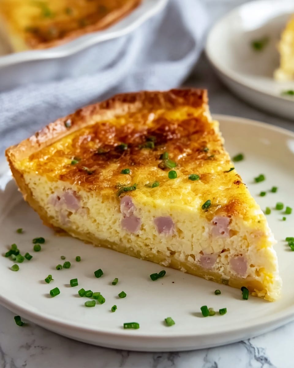 A slice of quiche is shown with three visible layers: a brown, flaky crust at the bottom; a thick, creamy yellow egg layer mixed with pieces of pink ham in the middle; and a golden-brown baked top sprinkled with small green chives. The slice sits on a white plate, placed on a white marbled surface, with additional chives scattered around the plate. The background is softly blurred with another plate of quiche visible. Photo taken with an iphone --ar 4:5 --v 7