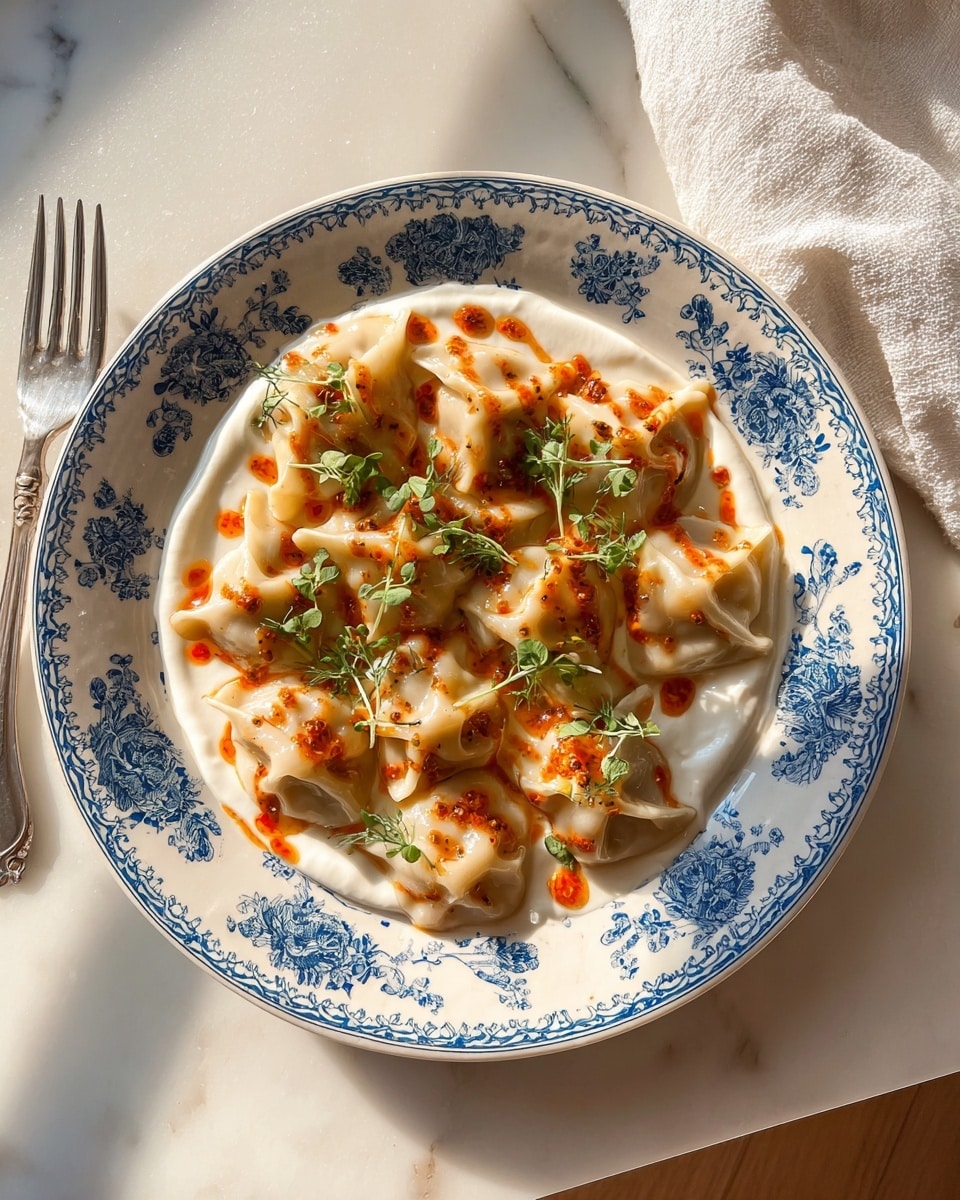 A white plate with blue floral patterns holds a single layer of dumplings arranged close together, each dumpling pale beige with a soft, slightly wrinkled texture. The dumplings sit on a thick base of creamy white yogurt spread evenly across the plate. A bright orange-red sauce is drizzled over the dumplings and yogurt in an irregular pattern, adding vibrant color. Small green herb sprigs are scattered lightly on top, providing a fresh contrast. The plate rests on a white marbled surface with natural light casting warm shadows. A gold fork and a white cloth napkin are partially visible near the plate. Photo taken with an iphone --ar 4:5 --v 7