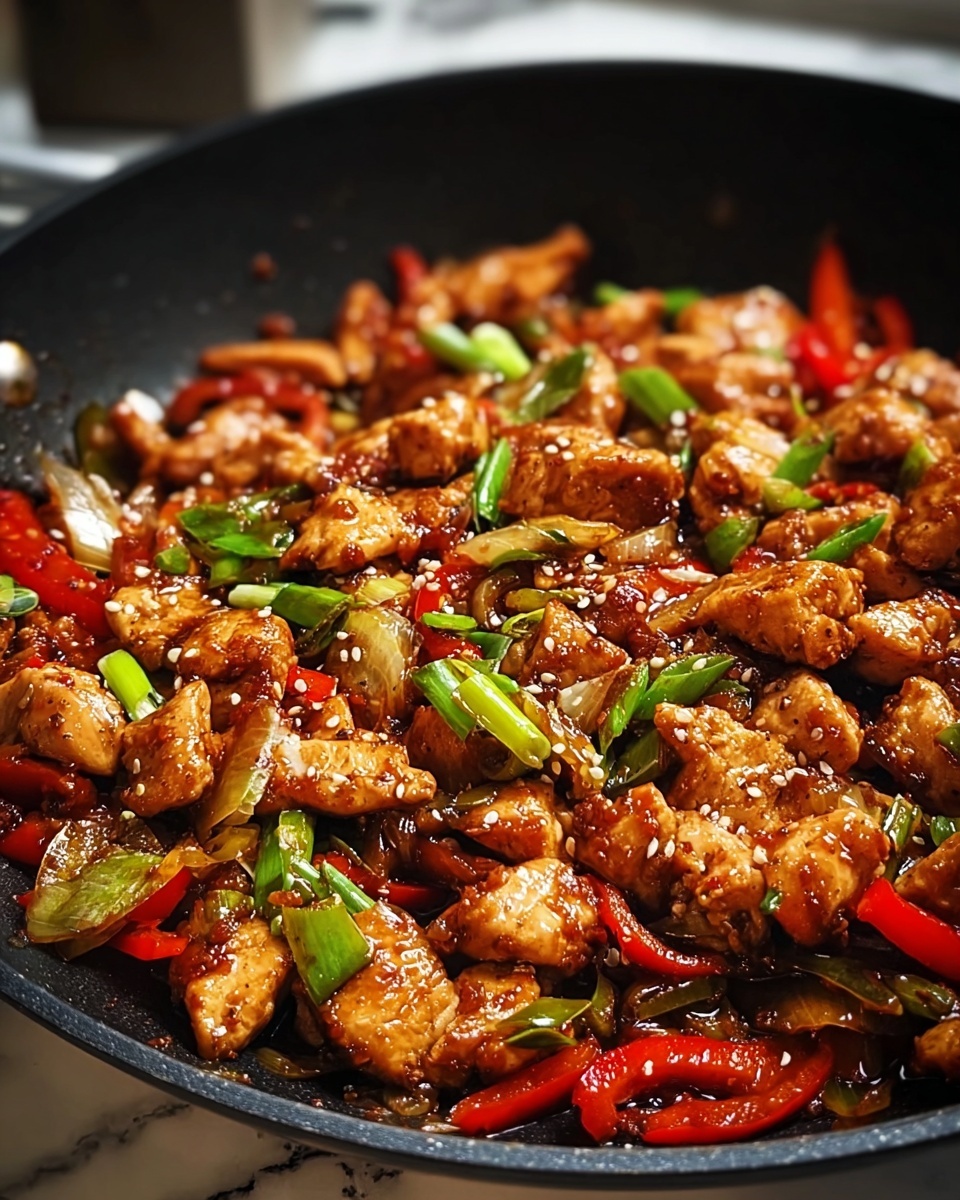 A close-up of a black frying pan filled with stir-fried chicken pieces covered in a shiny, reddish-brown sauce. The chicken is mixed with sliced red bell peppers, green onions, and light brown onion pieces. The dish is sprinkled with small white sesame seeds. The background shows a kitchen with a white marbled texture surface partially visible. photo taken with an iphone --ar 4:5 --v 7