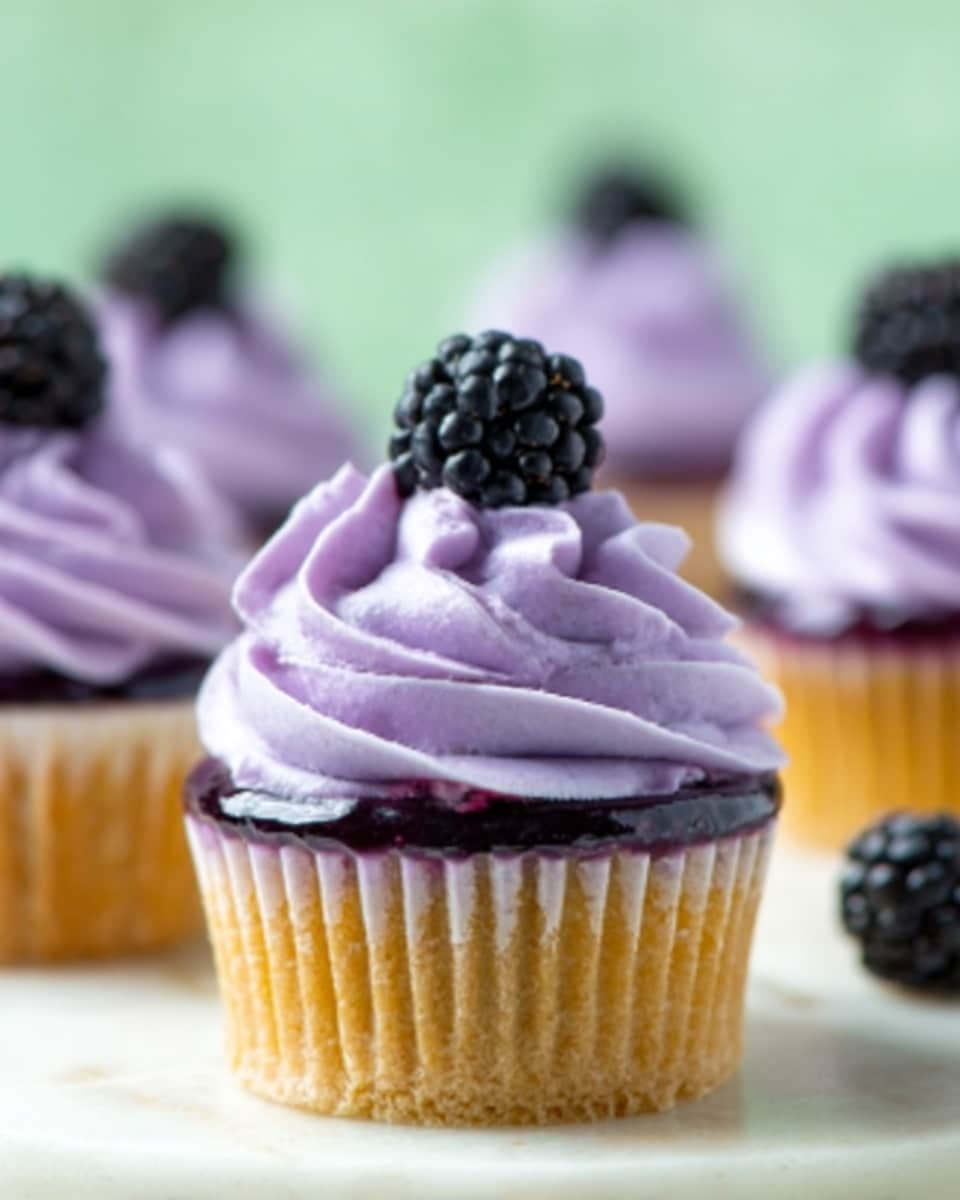 The image shows a close-up of a purple cupcake with three layers. The bottom layer is a purple cupcake base with a slightly rough texture. On top of this sits a smooth, dark purple jelly layer. Above the jelly is a swirl of light purple frosting with soft, smooth ridges. On the very top, there is a single fresh blackberry with a shiny, bumpy surface. The cupcake is placed on a white surface with a blurred green background. Photo taken with an iphone --ar 4:5 --v 7