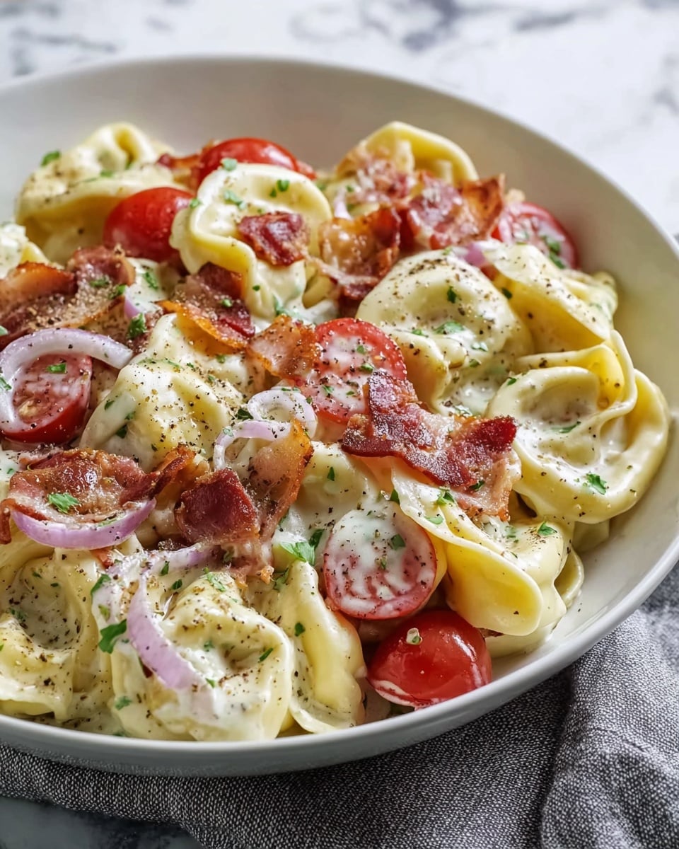 A white bowl is filled with about two layers of creamy tortellini pasta, each piece yellow with a soft texture, covered in a white sauce speckled with black pepper. Mixed in are halved, bright red cherry tomatoes and thin slices of red onion, adding fresh color. Crispy, brown bacon bits are scattered on top, along with chopped green herbs sprinkled unevenly, giving a fresh contrast. The bowl sits on a white marbled surface, showing the dish up close with clear detail. Photo taken with an iphone --ar 4:5 --v 7