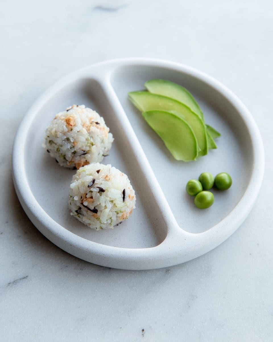 The image shows a gray divided plate with three sections placed on a white marbled surface. In the largest section, there are two rice balls mixed with small black and green bits, giving a speckled texture with white and light beige color. The middle section holds a light green smooth avocado slice, while the smallest section contains a few small green edamame pods. The background is bright and clean, highlighting the simple but colorful food arrangement. Photo taken with an iphone --ar 4:5 --v 7